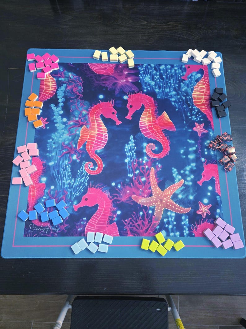 May include: A blue game mat featuring a vibrant underwater scene with seahorses, coral, and a starfish. Various colored game pieces surround the mat. The mat has a pink border and the text "Beach My Way".