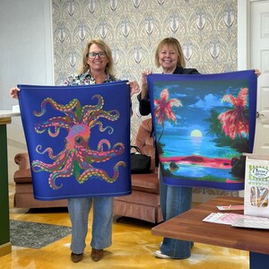 May include: Two women holding up blue fabric art. One features a colorful octopus design, the other a tropical sunset scene with palm trees. The fabric art is likely a scarf or wall hanging.