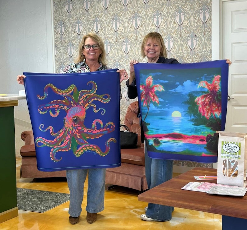 May include: Two women holding up blue fabric art. One features a colorful octopus design, the other a tropical sunset scene with palm trees. The fabric art is likely a scarf or wall hanging.