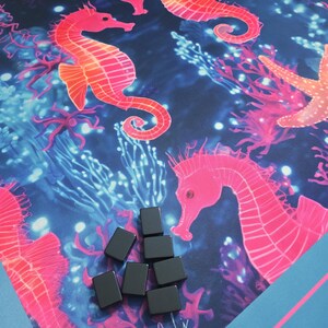 May include: A colorful game mat featuring a vibrant underwater scene with pink seahorses, coral, and starfish against a dark blue background. Several small, black rectangular game pieces are scattered on the mat. The mat has a blue border.