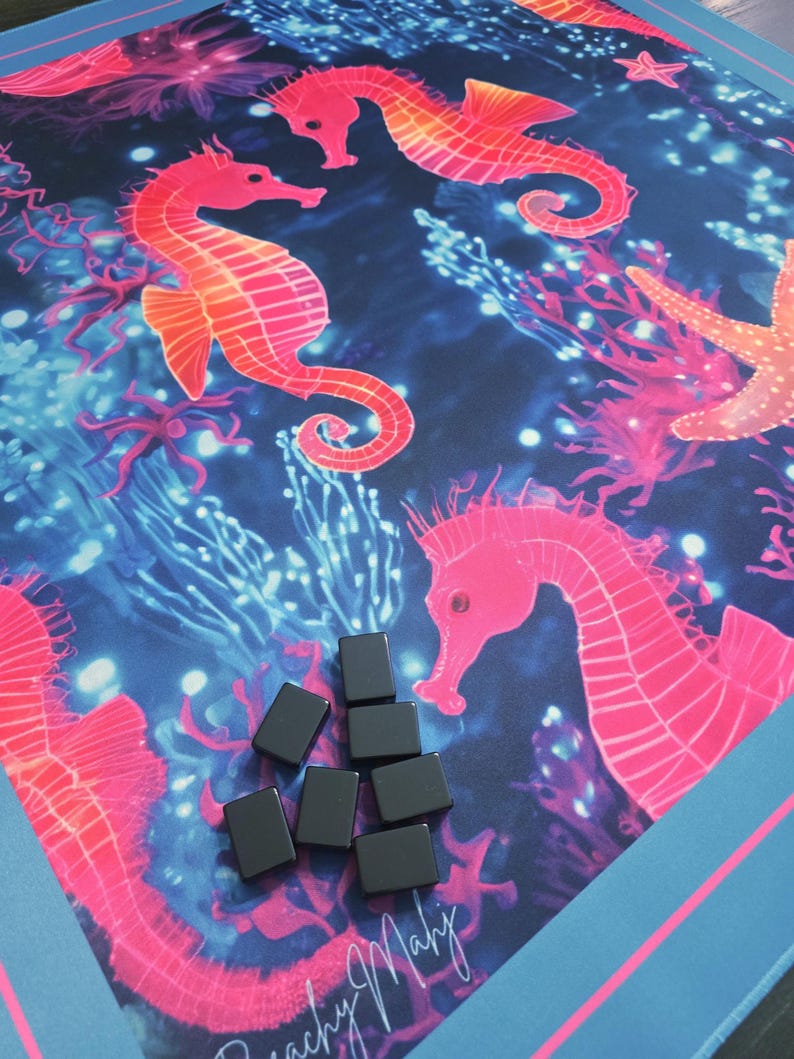 May include: A colorful game mat featuring a vibrant underwater scene with pink seahorses, coral, and starfish against a dark blue background. Several small, black rectangular game pieces are scattered on the mat. The mat has a blue border.