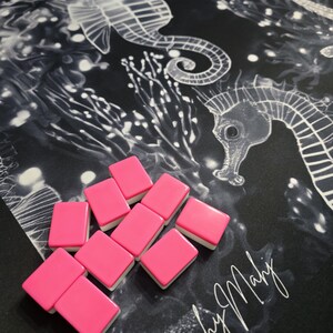 May include: A collection of pink rectangular tiles with white edges are arranged on a black surface. The surface features a white seahorse and coral design. The words "Beachy Mahy" are written in white script.