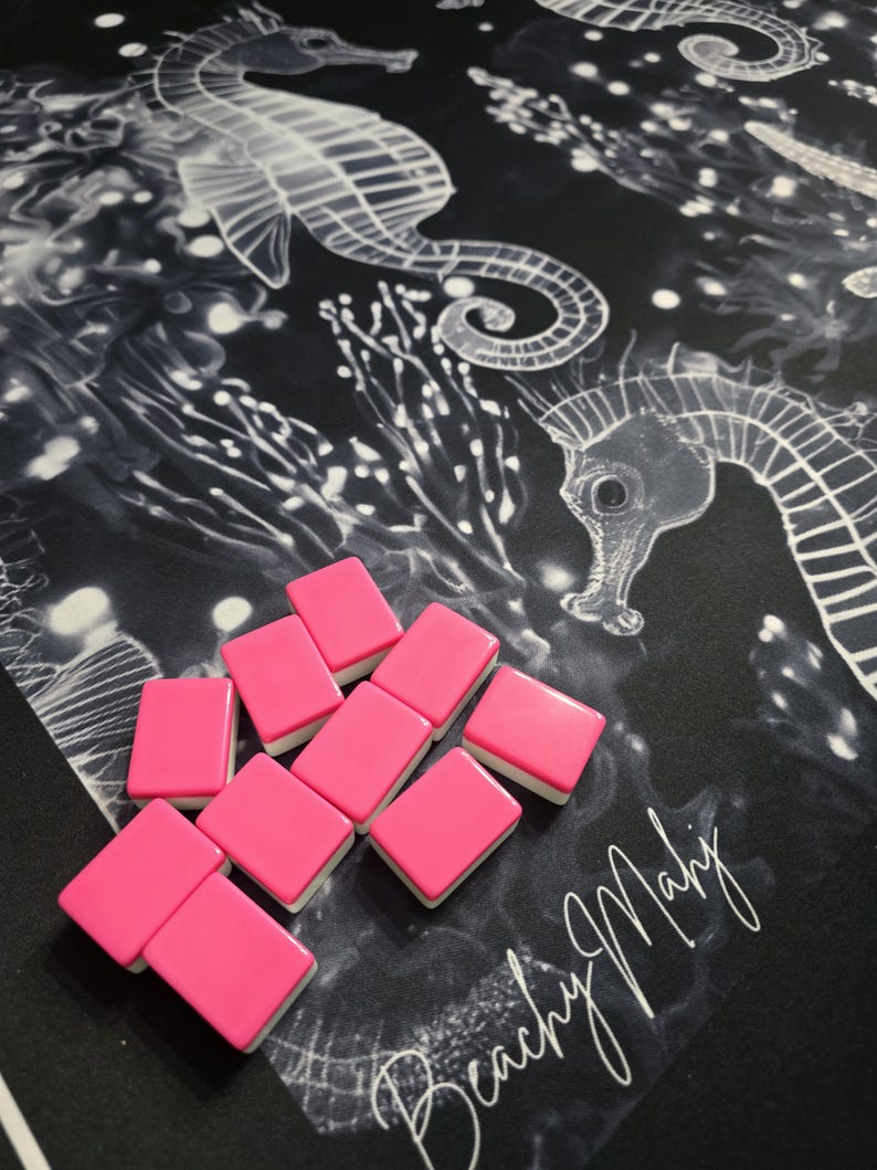 May include: A collection of pink rectangular tiles with white edges are arranged on a black surface. The surface features a white seahorse and coral design. The words "Beachy Mahy" are written in white script.