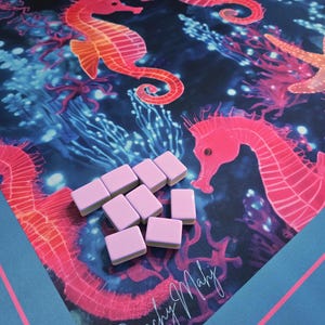 May include: A colorful game mat featuring a vibrant underwater scene with seahorses, coral, and starfish in shades of pink, orange, and blue. Light pink game tiles are scattered on the mat. The words "Beachy Mahj" are written in cursive.