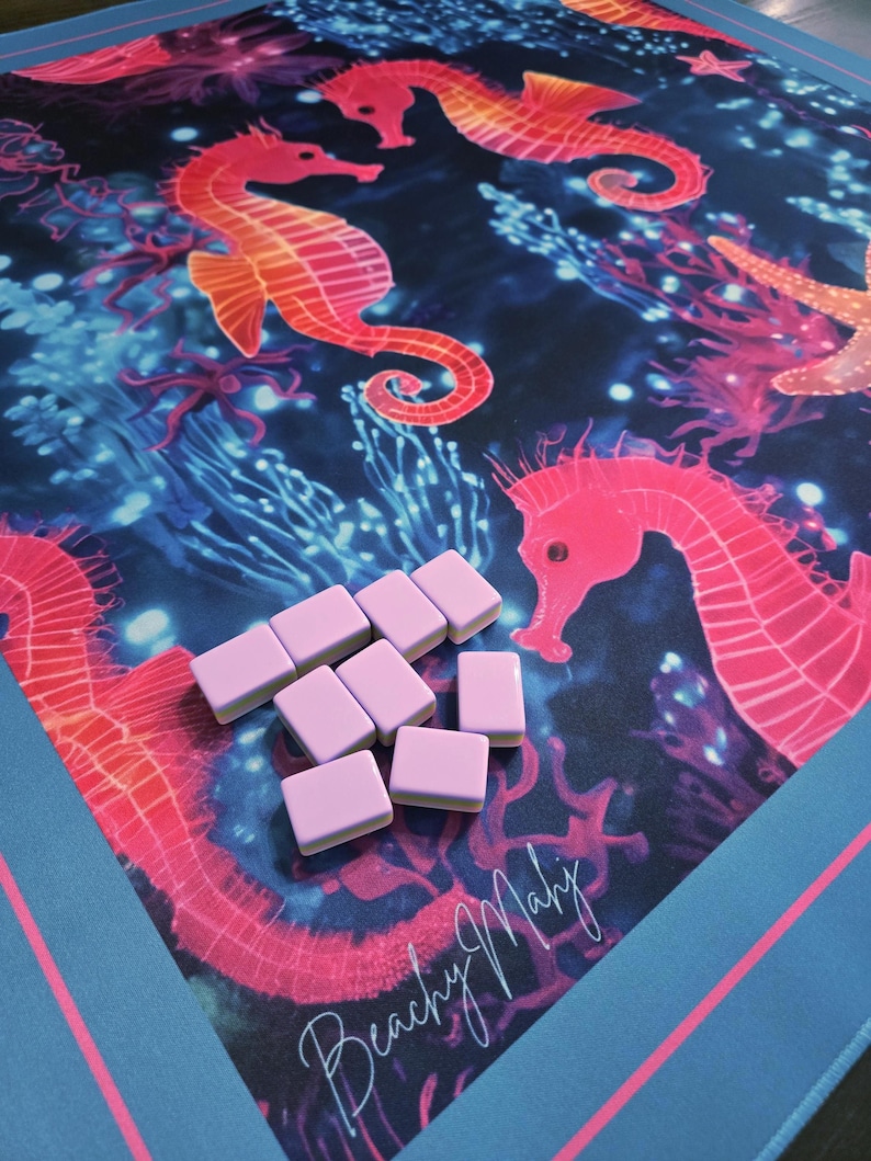 May include: A colorful game mat featuring a vibrant underwater scene with seahorses, coral, and starfish in shades of pink, orange, and blue. Light pink game tiles are scattered on the mat. The words "Beachy Mahj" are written in cursive.
