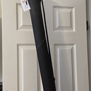 May include: A black, cylindrical carrying case with a shoulder strap hangs from a hook on a white door. The case is closed at the top with a drawstring. A small tag is attached to the case.