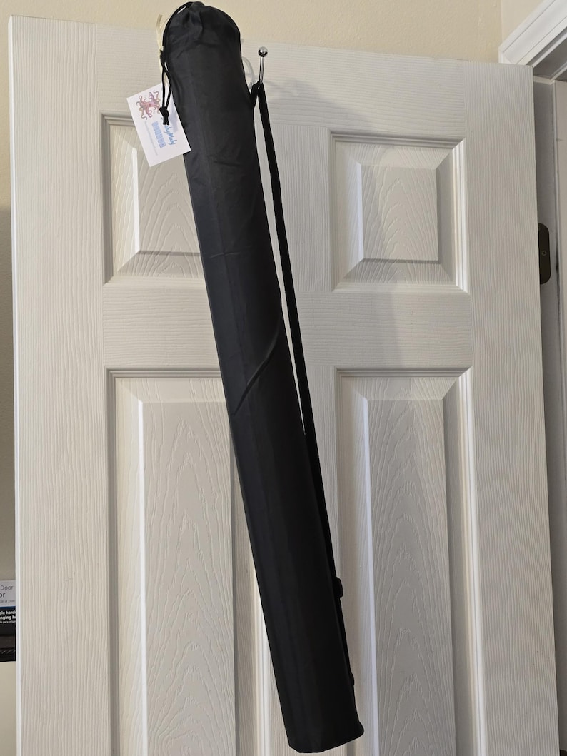 May include: A black, cylindrical carrying case with a shoulder strap hangs from a hook on a white door. The case is closed at the top with a drawstring. A small tag is attached to the case.