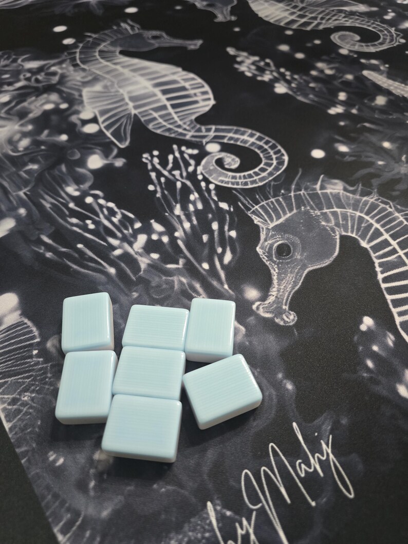 May include: Light blue square tiles arranged on a black and white seahorse patterned fabric. The fabric features a repeating design of seahorses and aquatic plants. The tiles are smooth and have a glossy finish.
