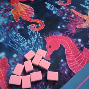May include: A pink Mahjong set with a blue and pink patterned mat featuring seahorses, coral, and starfish. The set includes pink rectangular tiles. The mat has the text "Beachy Mahj".