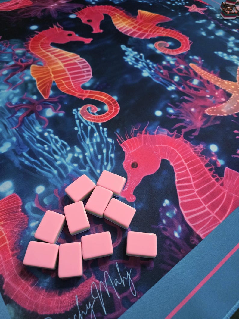 May include: A pink Mahjong set with a blue and pink patterned mat featuring seahorses, coral, and starfish. The set includes pink rectangular tiles. The mat has the text "Beachy Mahj".