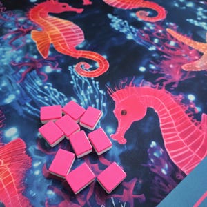 May include: A vibrant game mat featuring a marine design with seahorses in shades of orange and pink against a dark blue background. Pink game tiles with white edges are scattered on the mat. The mat has a blue border.