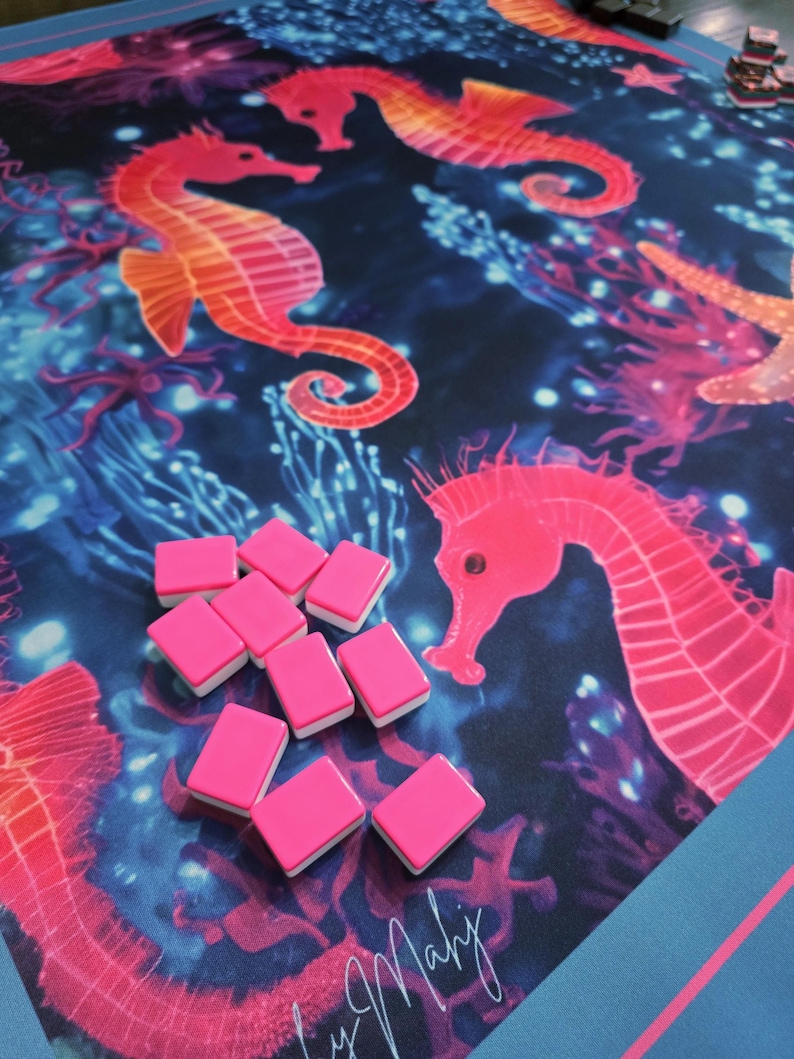 May include: A vibrant game mat featuring a marine design with seahorses in shades of orange and pink against a dark blue background. Pink game tiles with white edges are scattered on the mat. The mat has a blue border.
