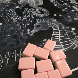 May include: Pink rectangular game tiles with rounded edges are scattered on a black surface. The surface has a white seahorse design and the cursive text "y Mah". The tiles are a solid pink color.