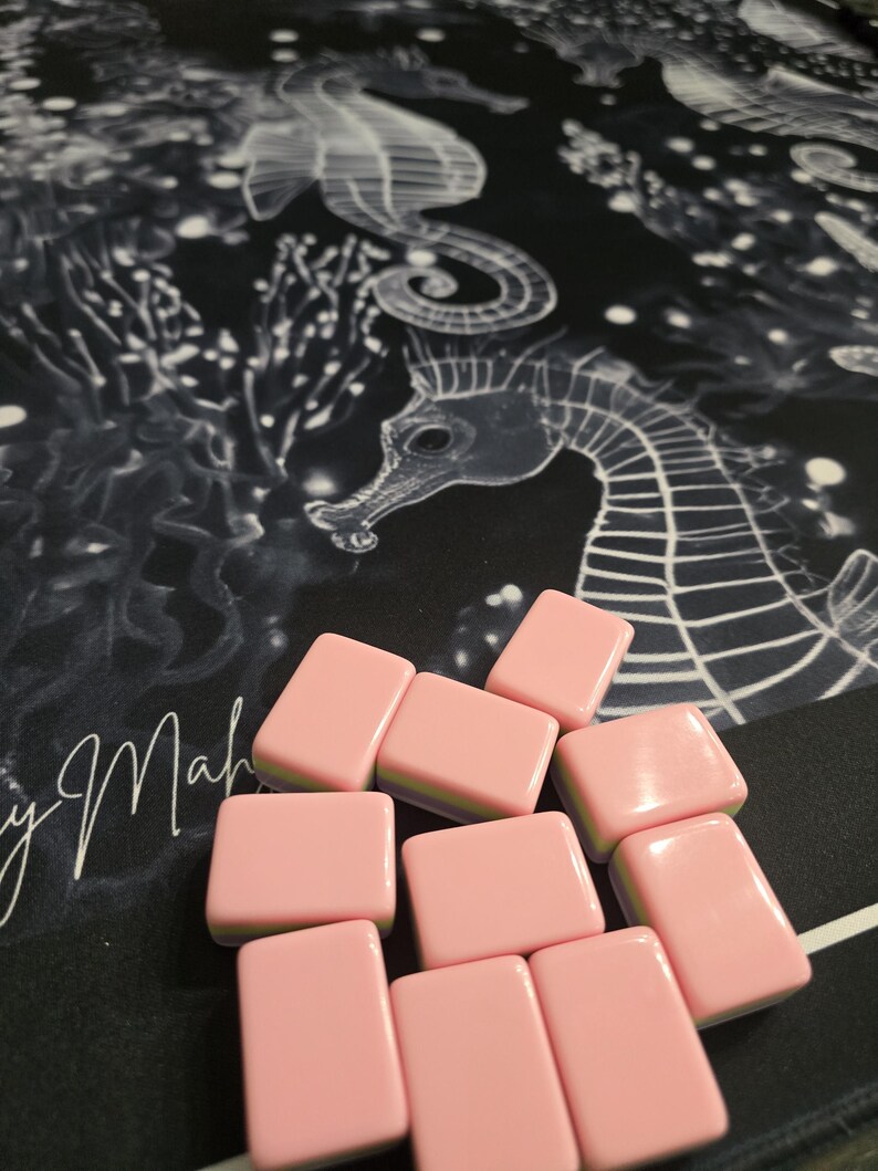 May include: Pink rectangular game tiles with rounded edges are scattered on a black surface. The surface has a white seahorse design and the cursive text "y Mah". The tiles are a solid pink color.