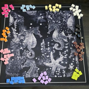 May include: A black board game mat featuring a white seahorse and starfish design. Various colored game pieces, including pink, blue, yellow, and orange, are scattered around the edges of the mat. The words "Beachy Maby" are printed on the bottom left.