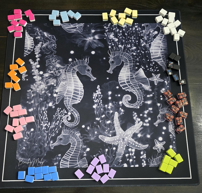 May include: A black board game mat featuring a white seahorse and starfish design. Various colored game pieces, including pink, blue, yellow, and orange, are scattered around the edges of the mat. The words "Beachy Maby" are printed on the bottom left.