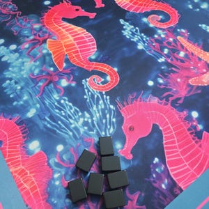 May include: A vibrant, ocean-themed game mat featuring coral, starfish, and seahorse illustrations in shades of pink and orange against a dark blue background. Several small, black rectangular game pieces are scattered on the mat. The mat has a blue border.