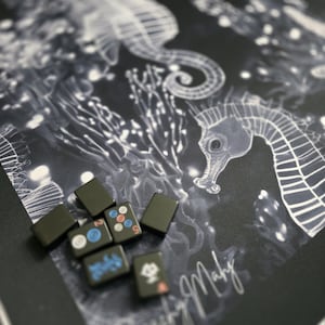 May include: A collection of black Mahjong tiles with colorful symbols and numbers, resting on a dark background with a seahorse and coral design. The design is in white and the tiles are rectangular.