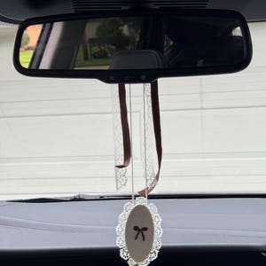 May include: An oval-shaped car hanging ornament featuring a beige center with a small dark brown bow graphic, surrounded by a white lace trim. The ornament is suspended by two thin brown ribbons and a white lace ribbon from a rearview mirror.