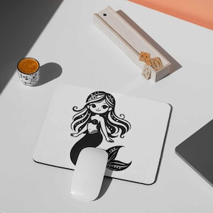 May include: A white mousepad featuring a black and white illustration of a mermaid. The mermaid has long, flowing hair and a detailed tail. A white computer mouse is positioned below the mousepad. A cup of coffee and a laptop are also visible.