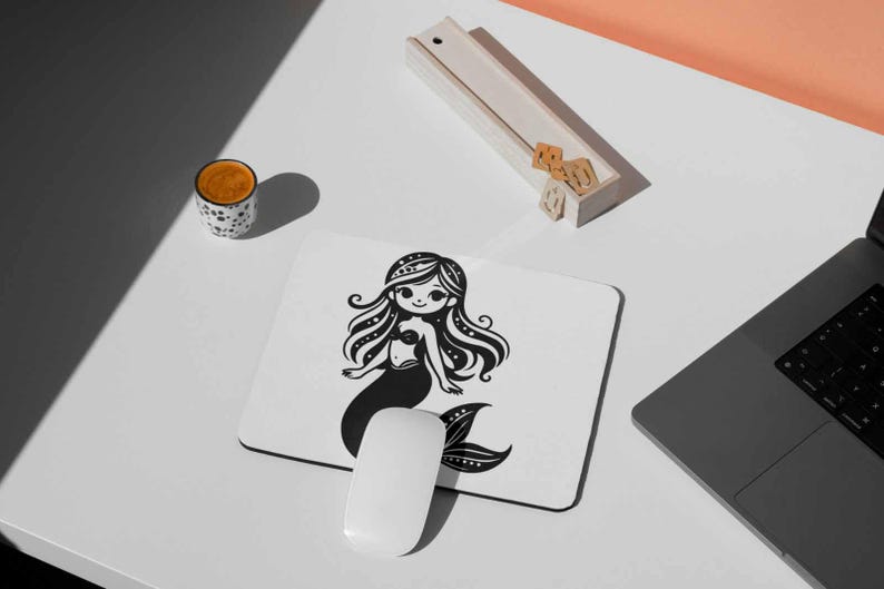 May include: A white mousepad featuring a black and white illustration of a mermaid. The mermaid has long, flowing hair and a detailed tail. A white computer mouse is positioned below the mousepad. A cup of coffee and a laptop are also visible.