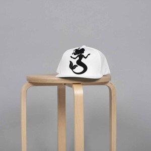 May include: A white baseball cap with a black silhouette of a mermaid. The cap is sitting on a light brown wooden stool against a gray background. The mermaid design features a flowing tail and long hair.