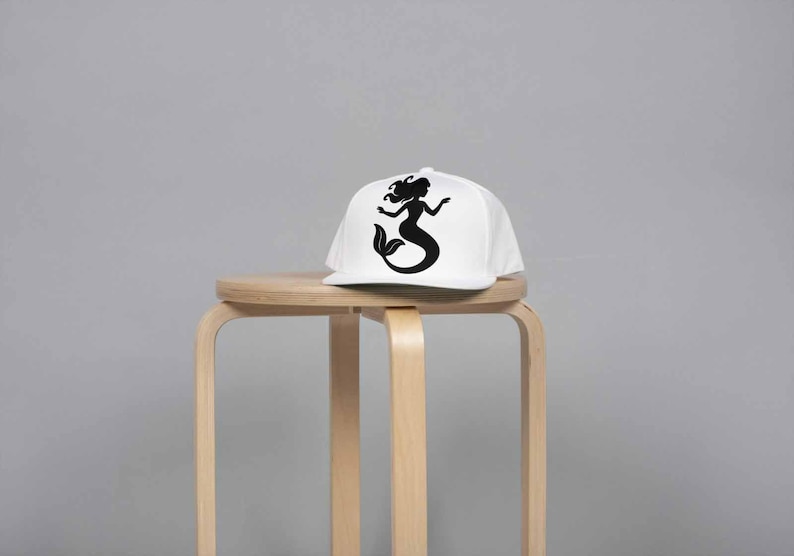May include: A white baseball cap with a black silhouette of a mermaid. The cap is sitting on a light brown wooden stool against a gray background. The mermaid design features a flowing tail and long hair.