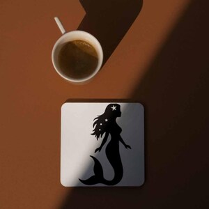 May include: A white square coaster with a black mermaid silhouette design, featuring stars in her hair, sits on a brown surface. A white coffee cup with dark liquid is positioned above the coaster. The image is lit with a shadow.