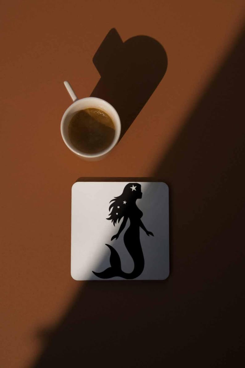 May include: A white square coaster with a black mermaid silhouette design, featuring stars in her hair, sits on a brown surface. A white coffee cup with dark liquid is positioned above the coaster. The image is lit with a shadow.