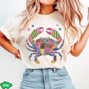 Puede incluir: Camiseta color crema con un diseño de cangrejo colorido. El cangrejo está compuesto por patrones de patchwork, incluyendo estampado de leopardo, cuadros y diseños florales. Las pinzas del cangrejo son rosas y amarillas a rayas, con detalles azules y amarillos. Estrellas azules y rosas rodean el cangrejo.