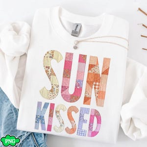 Könnte beinhalten: Weißes Sweatshirt mit den Worten "SUN KISSED" in bunten Patchwork-Buchstaben. Die Buchstaben bestehen aus verschiedenen floralen und gemusterten Stoffen in Rosa-, Orange-, Blau- und Gelbtönen. Eine silberne Halskette mit kleinem Anhänger liegt auf dem Sweatshirt.