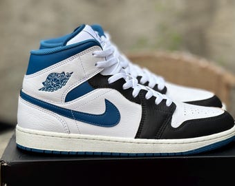 May include: White, black, and blue Air Jordan 1 Mid sneakers. The high-top shoes feature a white leather upper with blue accents on the collar, Swoosh, and outsole. The iconic Air Jordan logo is visible on the side.