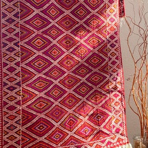 Könnte beinhalten: Ein lebendiger, handgewebter Teppich mit einem sich wiederholenden Rautenmuster in Rot-, Rosa- und Orangetönen. Das Design wird von einem weißen Rand umrahmt und mit Fransen versehen. Der Teppich ist wahrscheinlich aus Wolle oder einer ähnlichen Naturfaser gefertigt.