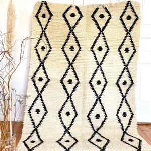 Könnte beinhalten: Cremefarbener, strukturierter Teppich mit einem sich wiederholenden schwarzen Rautenmuster. Der Teppich hat einen Fransenbesatz am unteren Rand und wird vor einer weißen Wand und einem Holzboden präsentiert. Ein dekorativer Einrichtungsgegenstand.