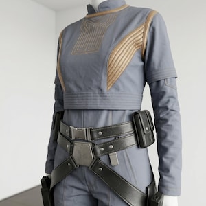 Puede incluir: Un atuendo de estilo futurista en gris con detalles dorados. La parte superior tiene costuras y ribetes dorados, y el atuendo incluye un cintur&oacute;n negro con bolsas y fundas. El maniqu&iacute; lleva un atuendo completo, incluyendo pantalones.