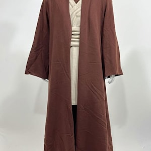 May include: A brown hooded robe with long sleeves, layered over a beige tunic and pants. The costume is displayed on a mannequin. The robe has a wide, flowing design, and the tunic has a V-neck. The outfit is a cosplay costume.