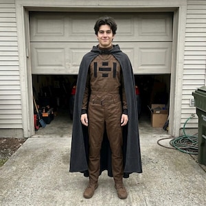 May include: A person wearing a brown jumpsuit with a black emblem on the chest, a black turtleneck, and brown boots. A dark gray cape is draped over the shoulders. The person is standing in front of a garage.