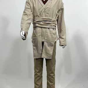 May include: A tan-colored costume displayed on a mannequin. The costume includes a long-sleeved tunic with a wrap-around belt, a brown undershirt, and matching pants. The outfit is designed with a layered appearance and a wide belt.