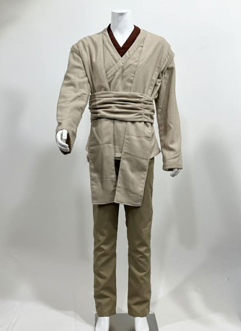 May include: A tan-colored costume displayed on a mannequin. The costume includes a long-sleeved tunic with a wrap-around belt, a brown undershirt, and matching pants. The outfit is designed with a layered appearance and a wide belt.