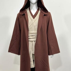 May include: A brown hooded robe with wide sleeves, layered over a cream-colored tunic and sash. The costume is displayed on a mannequin. The outfit is reminiscent of a Jedi or similar character.