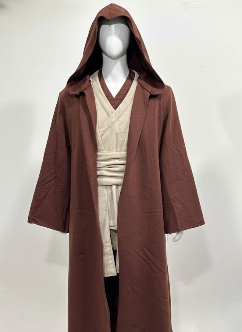 May include: A brown hooded robe with wide sleeves, layered over a cream-colored tunic and sash. The costume is displayed on a mannequin. The outfit is reminiscent of a Jedi or similar character.