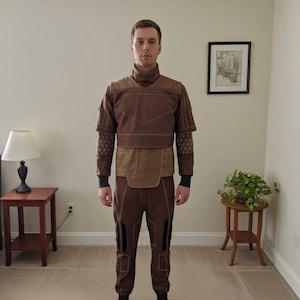 May include: Brown two-piece outfit with a high collar and geometric accents. The top has short sleeves and a contrasting panel at the waist. The pants have knee panels. The outfit is worn with black socks.