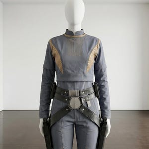 Puede incluir: Mono gris con detalles dorados y cintur&oacute;n negro con hebilla plateada. El conjunto incluye una parte superior de manga larga y pantalones, con fundas para dos pistolas negras. El maniqu&iacute; est&aacute; en una habitaci&oacute;n blanca.