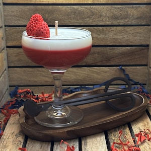 May include: A cocktail-shaped candle with a red and white layered design, topped with a strawberry. The candle sits in a clear glass on a wooden tray, with decorative accents.