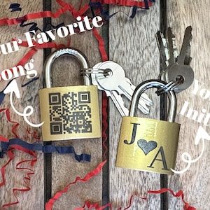 May include: Two gold padlocks with keys, one featuring a QR code and the other with initials "J A" and a heart symbol. Red, white, and blue streamers are scattered around the locks on a wooden surface.