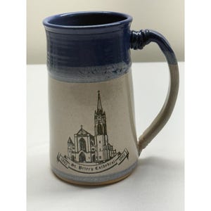 May include: A tall ceramic mug with a dark blue glazed rim and handle, transitioning to a light beige body. The front features a black line drawing of St. Peter's Cathedral with the text 1842 St. Peter's Cathedral 1992 printed on a banner.