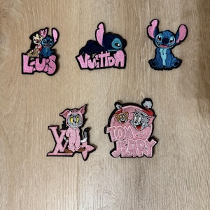 May include: A collection of five embroidered patches featuring cartoon characters and logos. The patches include Stitch, Tom and Jerry, and stylized text in pink and black. The patches are designed for application on clothing or accessories.