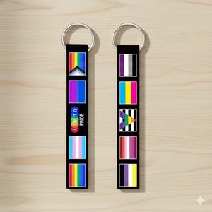 May include: Two black keychains with silver rings. Each keychain features a series of colorful LGBTQ+ pride flags. One keychain has the text "LGBTQ PRIDE" in a rainbow color scheme. The keychains are displayed on a light wood surface.