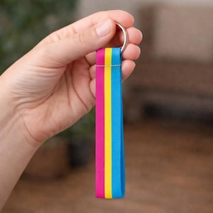 May include: A keychain with a colorful strap held by a hand. The strap features vertical stripes in pink, yellow, and blue, attached to a silver metal ring. The background is blurred, focusing attention on the keychain.