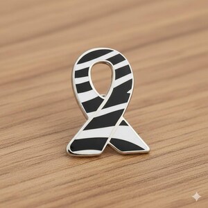 May include: A silver-toned ribbon pin with a black and white striped design. The pin is in the shape of a ribbon, with a polished finish. The pin is set against a wooden surface.